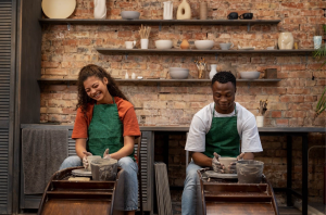 A mixed race lady in an orange tshirt and green apron and a black man in a white tshirt and green apron sat side by side in a pottery studio spinning clay on a wheel and smiling. 